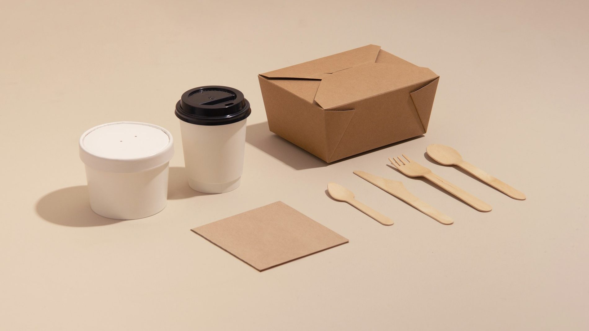 Caja kraft, vaso con tapa, pote de sopa y cubiertos de madera sobre fondo beige